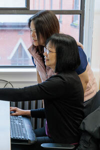 One woman stands behind another woman seated at a desk, pointing to a computer screen