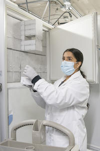 A woman in a lab coat wearing a mask stands in front of a freezer holding a vial