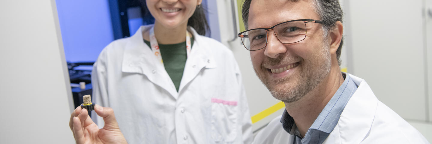 A man wearing a lab coat holds an item in his hand in the foreground while a smiling woman stands behind him