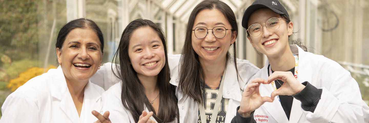 Four women in lab coats smile at the camera with their hands in heart shapes