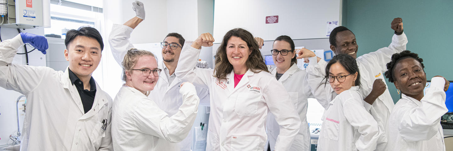A group of people in lab coats stand in a lab with their arms raised to indicate strength