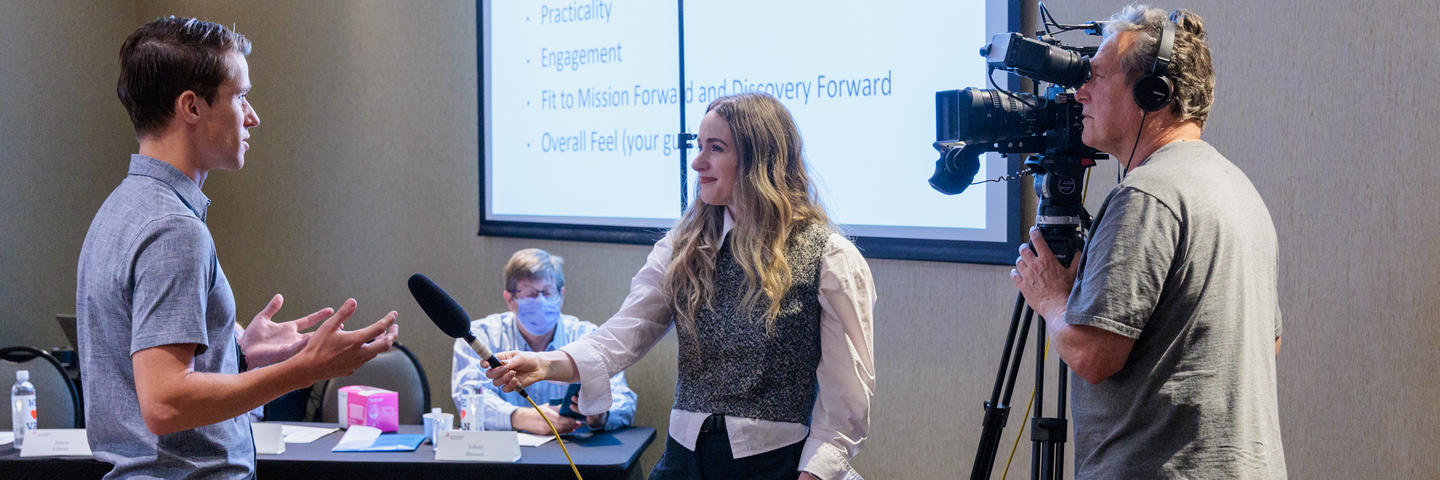 A man and a woman stand in front of a projector screen while she holds a microphone for him to speak into