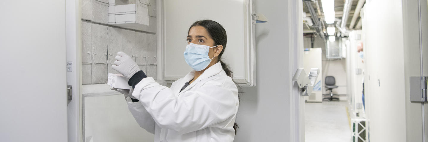 A woman in a lab coat wearing a mask stands in front of a freezer holding a vial
