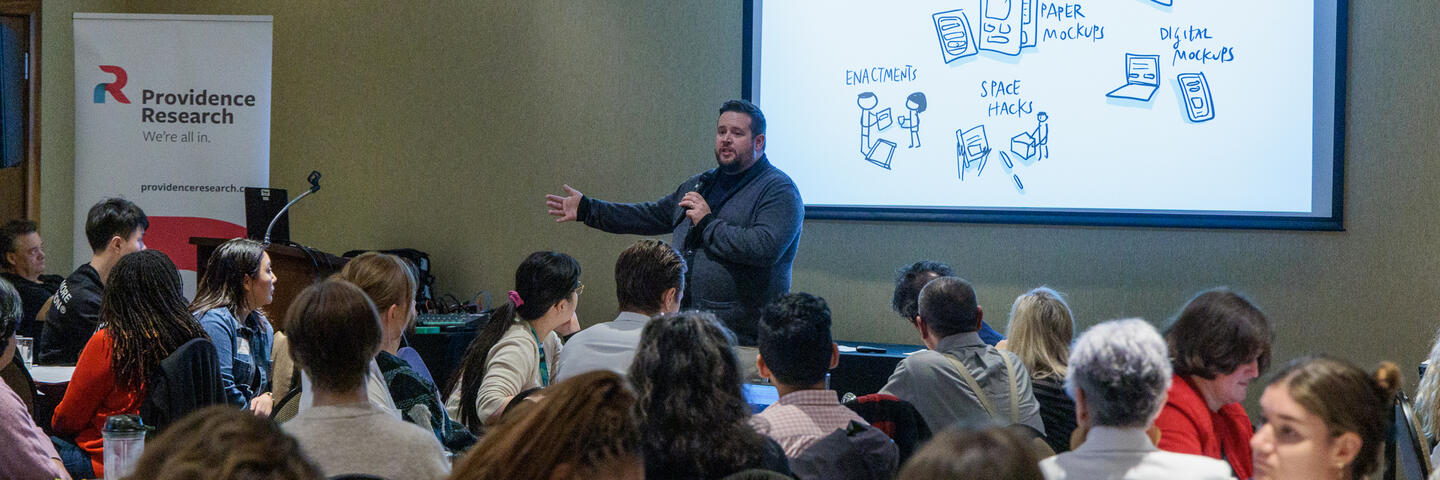 A man stands at the front of a room of people in front of a projector screen