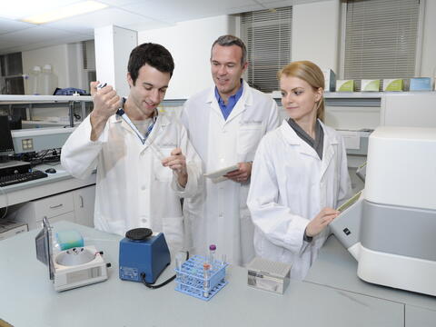 Three people in lab coats stand in a lab looking at a test tube